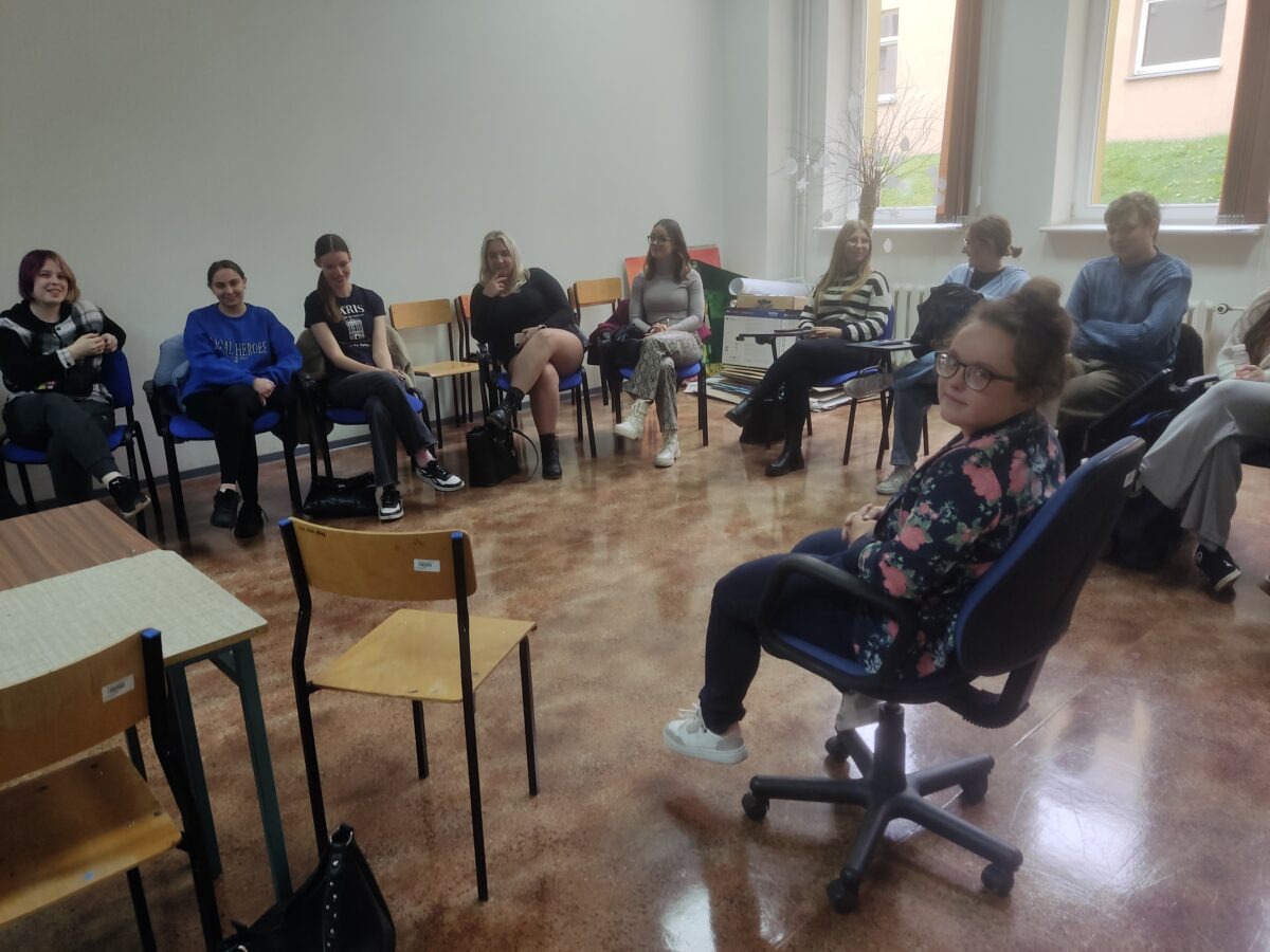 Sala wykładowa. Na pierwszym planie zwrócona do kadru siedząca na krześle kobieta. Dookoła niej na krzesłach siedzą studenci. Po prawej stronie są okna.