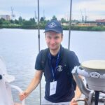 zdjęcie przestawiające chodzącego młodego chłopaka po statku w tle woda oraz w oddali infrastruktura