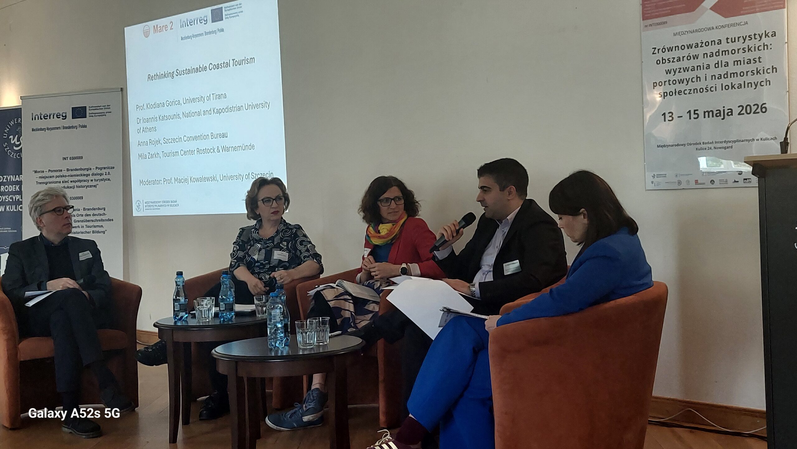 Panel dyskusyjny podczas konferencji naukowej. Pięć osób siedzi półkolem w pomarańczowych fotelach, przed nimi stoją dwa okrągłe stoliki z butelkami wody i szklankami. Po lewej stronie siedzi mężczyzna o jasnych włosach w ciemnym garniturze z założoną nogą. Obok niego znajduje się kobieta o jasnej karnacji, w okularach, z krótkimi włosami i w granatowo‑białej sukience w abstrakcyjny wzór. Trzecia osoba to kobieta o średniej długości ciemnych włosach, w czerwonej marynarce i kolorowej chuście, trzyma notatki na kolanach. Kolejna osoba, mężczyzna o ciemnych włosach i jasnej karnacji w ciemnej marynarce, trzyma mikrofon i mówi; na kolanach ma biały plik dokumentów. Po prawej siedzi kobieta o jasnej karnacji w niebieskim garniturze, pochylona w jego stronę. Na ścianie za panelistami z lewej strony stoi pionowy baner projektu Interreg z logotypami Unii Europejskiej i partnerów. Obok, na środku, jest duży ekran z prezentacją; widoczny jest tytuł po angielsku „Rethinking Sustainable Coastal Tourism” oraz lista prelegentów i moderatora, w tym m.in. University of Tirana, National and Kapodistrian University of Athens, Szczecin Convention Bureau, Tourism Center Rostock & Warnemünde oraz University of Szczecin. Po prawej stronie na ścianie wisi plakat konferencji po polsku z tytułem „Zrównoważona turystyka obszarów nadmorskich: wyzwania dla miast portowych i nadmorskich społeczności lokalnych” oraz terminem „13–15 maja 2026”. Na dole plakatu znajdują się logotypy instytucji współorganizujących. W prawym dolnym rogu zdjęcia fragment mównicy oraz kable na podłodze.