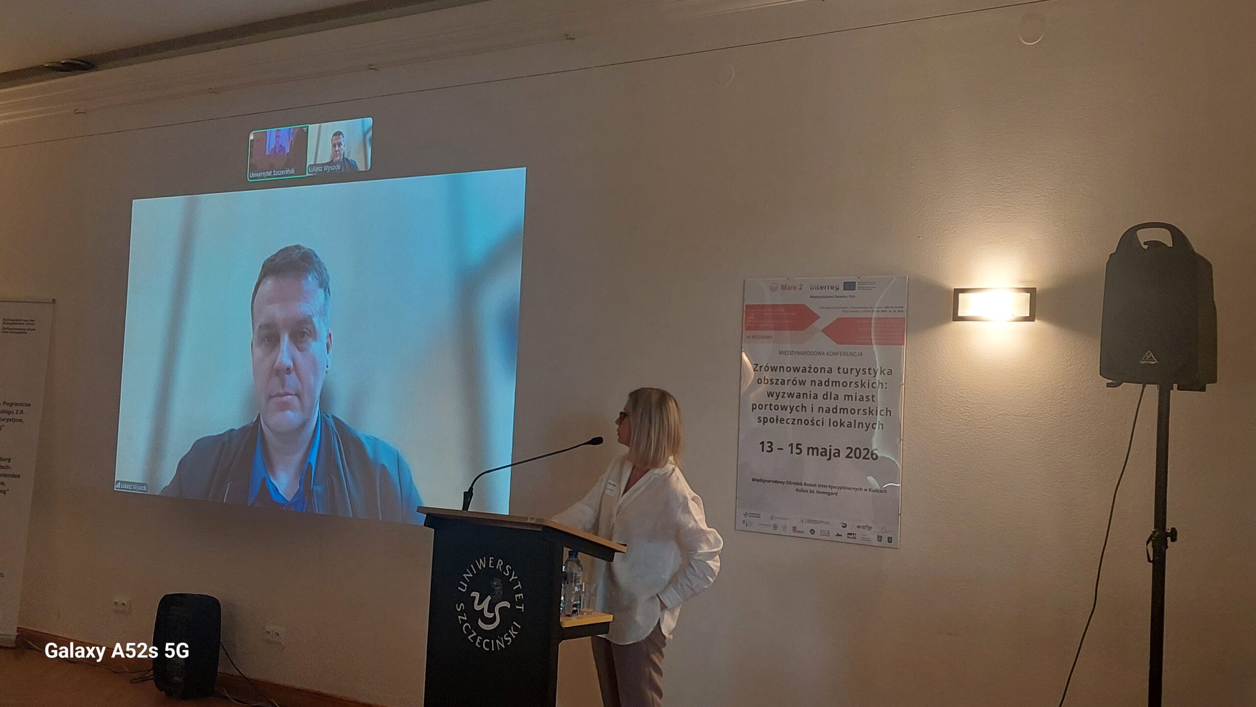 Duży ekran projekcyjny z twarzą mężczyzny podczas wideokonferencji w sali wykładowej. Na ekranie wyświetlany jest mężczyzna o jasnej karnacji i krótkich włosach, ubrany w niebieską koszulę i ciemną kurtkę lub marynarkę. Nad jego obrazem widać dwa małe okienka z innymi uczestnikami połączenia. Po prawej stronie stoi mównica z logo Uniwersytetu Szczecińskiego; za mównicą znajduje się kobieta o jasnej karnacji i blond, prostych włosach do ramion, ubrana w jasną koszulę i spodnie. Kobieta stoi bokiem do ekranu i zwrócona jest w stronę publiczności; na mównicy leży mikrofon i stoi butelka wody. Na ścianie po prawej stronie wisi jasny plakat konferencji z czerwonym nagłówkiem. Tekst na plakacie (w miarę czytelny) brzmi: „Między Morzem a Interesem Zrównoważona turystyka obszarów nadmorskich: wyzwania dla miast portowych i nadmorskich społeczności lokalnych 13 – 15 maja 2026” Niżej są drobniejsze, słabo czytelne logotypy i napisy partnerów. Obok plakatu, po prawej, znajduje się głośnik na wysokim statywie, z podłączonym kablem. Na ścianie świeci prostokątna kinkietowa lampa, dająca ciepłe światło.