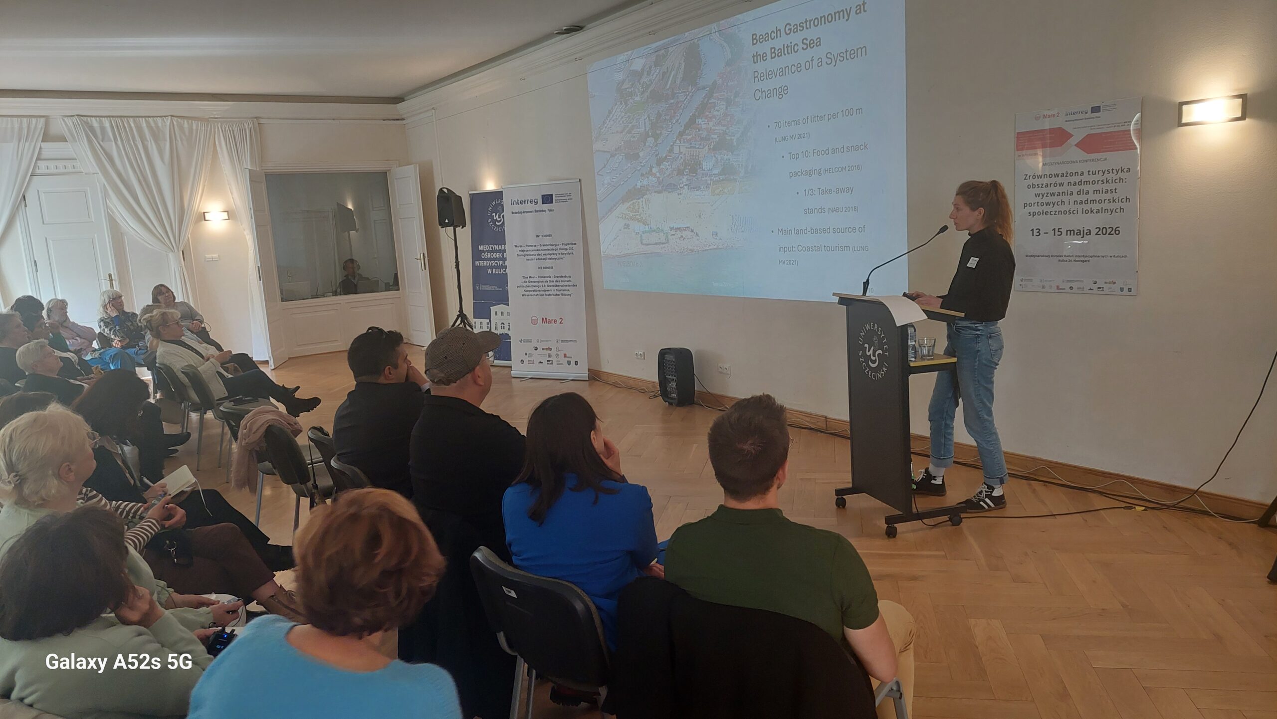 Sala konferencyjna z prezentacją multimedialną. Po prawej stronie stoi przy mównicy młoda kobieta o jasnej skórze, w czarnym golfie i jasnych dżinsach, w sportowych butach. Mówi do mikrofonu i patrzy w stronę dużego ekranu z rzutnika. Na ekranie widać slajd po angielsku zatytułowany „Beach Gastronomy at the Baltic Sea – Relevance of a System Change”; pod tytułem są akapity tekstu i zdjęcie fragmentu wybrzeża. Przed mównicą siedzi publiczność na krzesłach w kilku rzędach, około dwudziestu kilku osób, głównie dorośli i seniorzy, skierowani twarzami w stronę prelegentki. Część osób trzyma notatki lub telefony. Za prelegentką, bliżej środka ściany, stoją dwie wysokie, pionowe plansze–banery z logotypami, tekstem i znakami Unii Europejskiej oraz programu Interreg (szczegółowy tekst jest zbyt mały, by go odczytać). Po prawej stronie ściany wisi kolejny plakat z datą „13 – 15 maja 2026” i informacją o zrównoważonej turystyce obszarów nadmorskich; pozostały tekst jest drobny i nieczytelny. Pomieszczenie ma jasne ściany, drewnianą podłogę i po lewej stronie duże, zasłonięte zasłonami okna lub przeszklone drzwi.