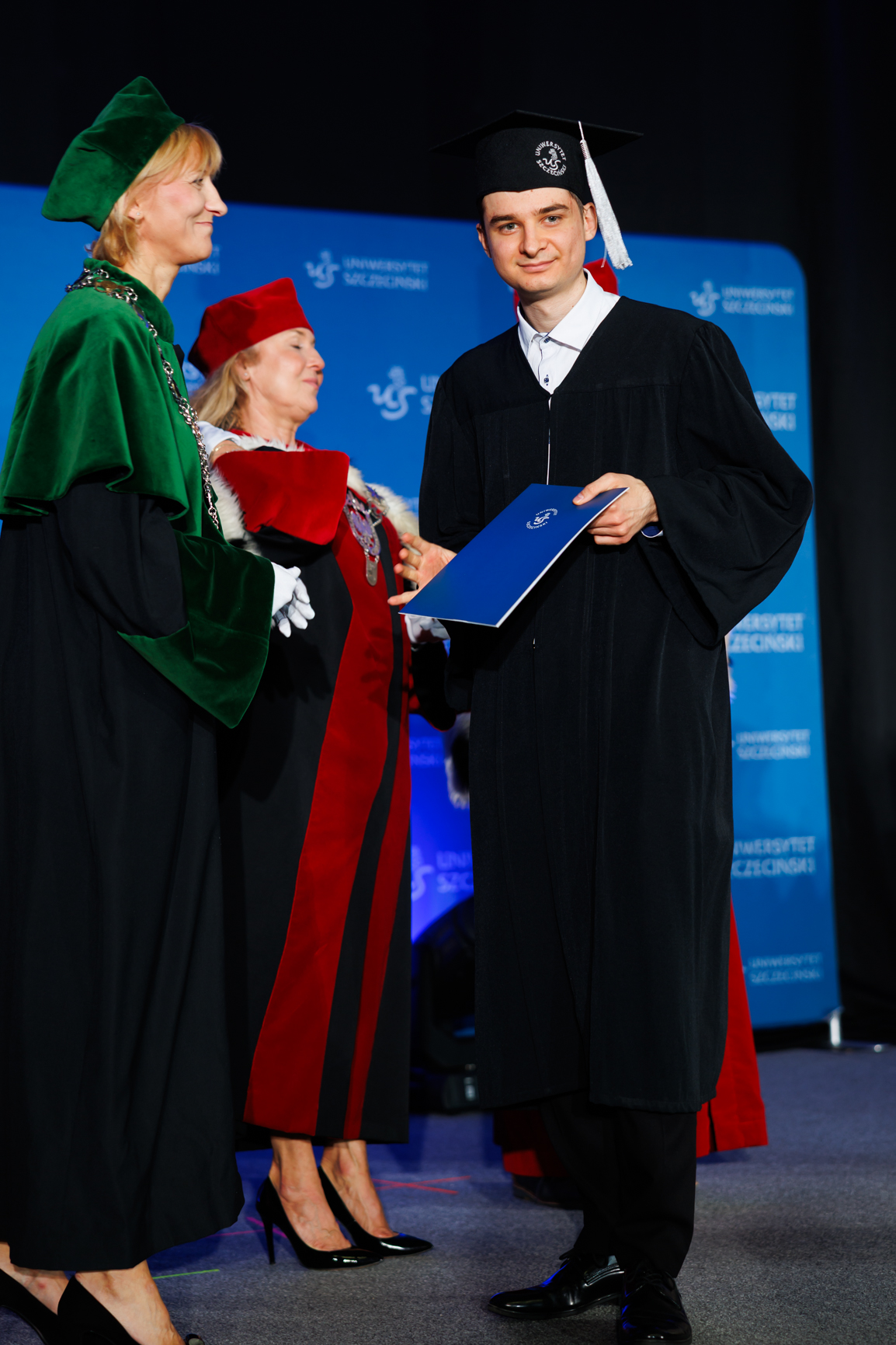 Młody mężczyzna w todze i birecie podczas ceremonii rozdania dyplomów. Stoi po prawej stronie, ma jasną karnację, ciemne krótkie włosy i lekki uśmiech. Na głowie ma czarny biret z białym chwostem i okrągłą naszywką z godłem uczelni. Ubrany jest w długą czarną togę, pod spodem widać biały kołnierzyk koszuli. W obu dłoniach trzyma niebieską teczkę lub dyplom z białym logo uczelni na środku. Po lewej stronie stoi kobieta w uroczystej czarno‑zielonej todze; ma jasną skórę, blond włosy do szyi i zielony aksamitny biret. Na szyi widoczny jest łańcuch rektorski. Za nią, lekko w tle, stoi kolejna kobieta w czarno‑czerwonej todze z białym futrzanym kołnierzem, czerwonym biretem i dużym łańcuchem z medalionem. W tle znajduje się niebieska ściana z powtarzającym się białym logo „Uniwersytet Szczeciński” i znakiem graficznym uczelni. Podłoga jest szara, a osoby mają na sobie eleganckie, czarne, lakierowane buty. Cała scena wygląda jak oficjalna uroczystość akademicka na scenie.