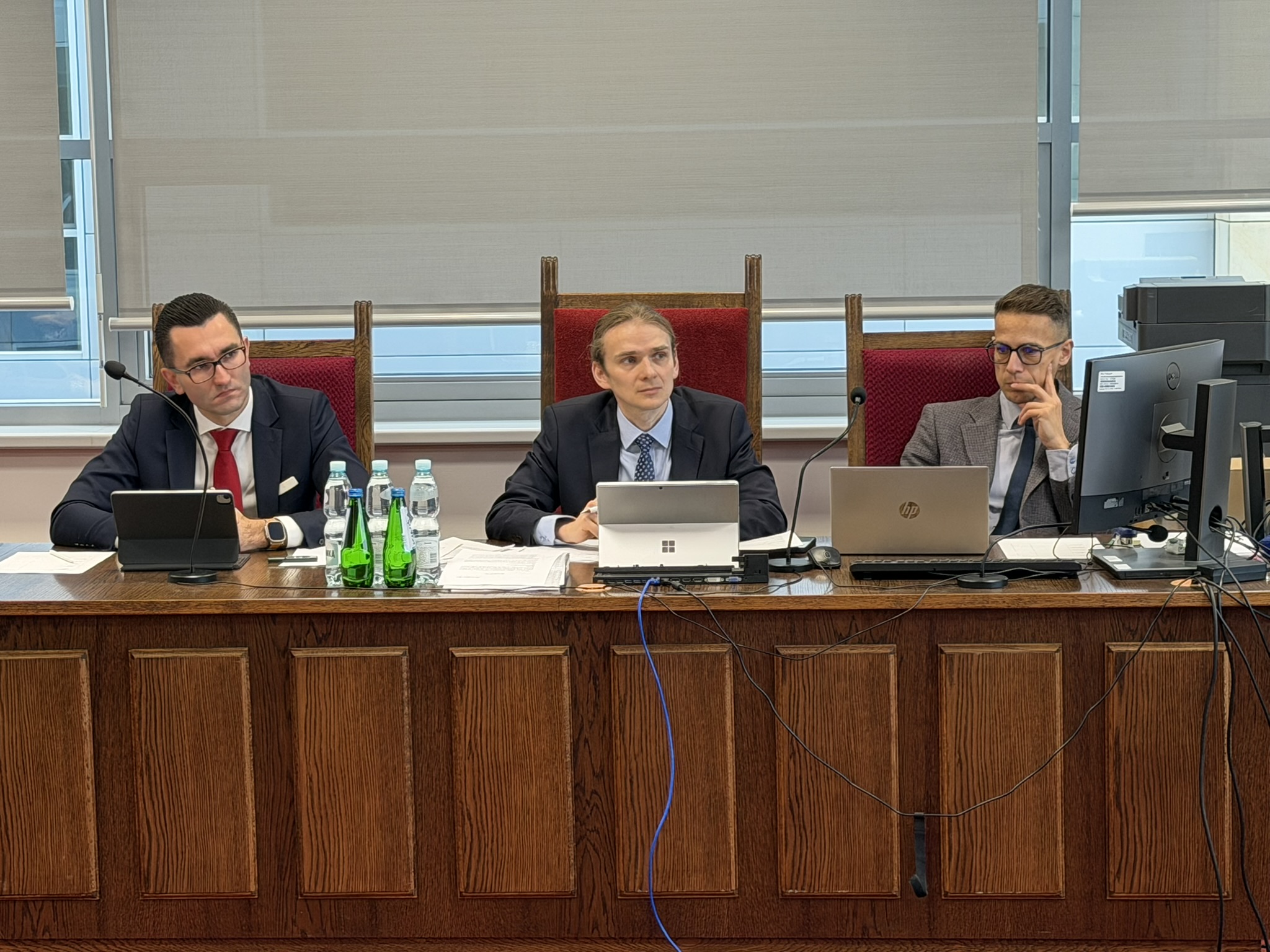 Pomieszczenie do symulacji rozprawy sądowej. Za drewnianymi zabudowanymi stołami, na krzesłach obitych bordowym materiałem, siedzi trzech mężczyzn (skład sędziowski) – przed nimi stoją laptopy lub tablety.