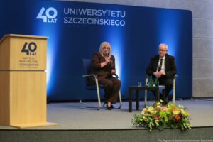 Konferencja podczas 40-lecia Instytutu Nauk o Polityce i Bezpieczeństwie Uniwersytetu Szczecińskiego. Na scenie dwoje rozmówców na krzesłach. Pośrodku czarny kwadratowy stolik. W tle granatowa ścianka z napisem: 40 lat Uniwersytetu Szczecińskiego.