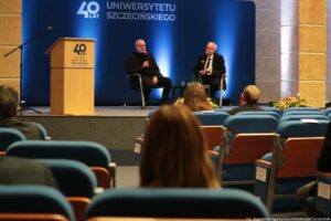 Konferencja podczas 40-lecia Instytutu Nauk o Polityce i Bezpieczeństwie Uniwersytetu Szczecińskiego. Aula. Ujęcie tyłem na słuchaczy. Na scenie dwóch rozmówców na krzesłach. Pośrodku czarny kwadratowy stolik. W tle granatowa ścianka z napisem: 40 lat Uniwersytetu Szczecińskiego.