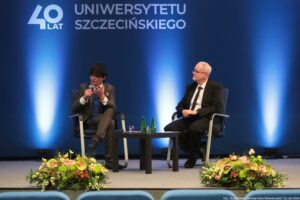 Konferencja podczas 40-lecia Instytutu Nauk o Polityce i Bezpieczeństwie Uniwersytetu Szczecińskiego. Na scenie dwóch rozmówców na krzesłach. Pośrodku czarny kwadratowy stolik. W tle granatowa ścianka z napisem: 40 lat Uniwersytetu Szczecińskiego.