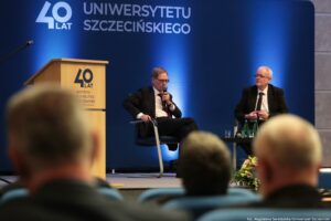Konferencja podczas 40-lecia Instytutu Nauk o Polityce i Bezpieczeństwie Uniwersytetu Szczecińskiego. Aula. Ujęcie tyłem na słuchaczy. Na scenie dwóch rozmówców na krzesłach. Pośrodku czarny kwadratowy stolik. W tle granatowa ścianka z napisem: 40 lat Uniwersytetu Szczecińskiego.