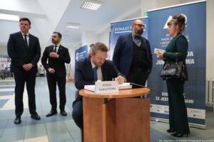 Uroczystość 40-lecia Instytutu Nauk o Polityce i Bezpieczeństwie Uniwersytetu Szczecińskiego. Na pierwszym planie mężczyzna wpisujący się do księgi pamiątkowej. W tle czworo ludzi.