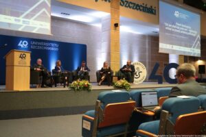 Konferencja podczas 40-lecia Instytutu Nauk o Polityce i Bezpieczeństwie Uniwersytetu Szczecińskiego. Aula. Ujęcie tyłem na słuchaczy. Na scenie sześcioro rozmówców na fotelach (2 kobiety, 4 mężczyzn). W tle granatowa ścianka z napisem: 40 lat Uniwersytetu Szczecińskiego. Po prawej ekran z tytułem dyskusji dotyczącej rozwoju polskiej politologii.
