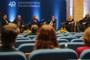 Konferencja podczas 40-lecia Instytutu Nauk o Polityce i Bezpieczeństwie Uniwersytetu Szczecińskiego. Aula. Ujęcie tyłem na słuchaczy. Na scenie sześcioro rozmówców na fotelach (2 kobiety, 4 mężczyzn). W tle granatowa ścianka z napisem: 40 lat Uniwersytetu Szczecińskiego.