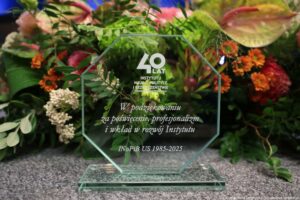 Szklana ośmiokątna statuetka z grawerowanym napisem: „40 lat Instytutu Nauk o Polityce i Bezpieczeństwie Uniwersytetu Szczecińskiego. W podziękowaniu za poświecenie, profesjonalizm i wkład w rozwój Instytutu. INoPiB US 1985-2025”. W tle kwiaty.