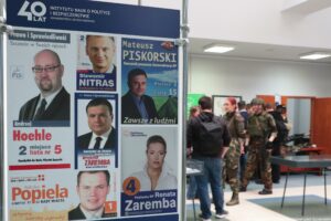 40-lecie Instytutu Nauk o Polityce i Bezpieczeństwie Uniwersytetu Szczecińskiego. Korytarz. Po lewej fragment wystawy plansz z plakatami i ulotkami wyborczymi absolwentów uczelni. W tle uczestnicy wydarzenia.
