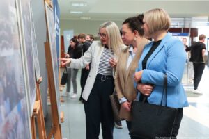 Trzy pracownice uczelni (po prawej – ujęcie bokiem) oglądają wystawę jubileuszową z okazji 40-lecia Instytutu Nauk o Polityce i Bezpieczeństwie Uniwersytetu Szczecińskiego. Plansze ze zdjęciami ustawione są na sztalugach. W tle inni uczestnicy wydarzenia.
