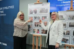 40-lecie Instytutu Nauk o Polityce i Bezpieczeństwie Uniwersytetu Szczecińskiego. Korytarz. Dwie przedstawicielki uczelni pokazują palcami wskazującymi na swoje zdjęcia archiwalne prezentowane na fotograficznej wystawie jubileuszowej.
