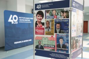 40-lecie Instytutu Nauk o Polityce i Bezpieczeństwie Uniwersytetu Szczecińskiego. Korytarz. Wystawa plansz z plakatami i ulotkami wyborczymi absolwentów uczelni. W tle granatowa ścianka z napisem: 40 lat Instytutu Nauk o Polityce i Bezpieczeństwie Uniwersytetu Szczecińskiego.