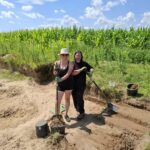 Dwie kobiety pozujące do zdjęcia podczas wykopalisk trzymające łopaty w rękach.