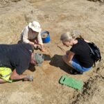 3 osoby wykonujące prace archeologiczne w otwartej przestrzeni.