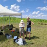 Dwie kobiety przyglądające się dokumentacji podczas wykopalisk, w tle błękitne niebo z kilkoma chmurkami, zasiane pole oraz rozstawiony namiot.