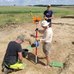 3 osoby wykonujące prace archeologiczne w otwartej przestrzeni.