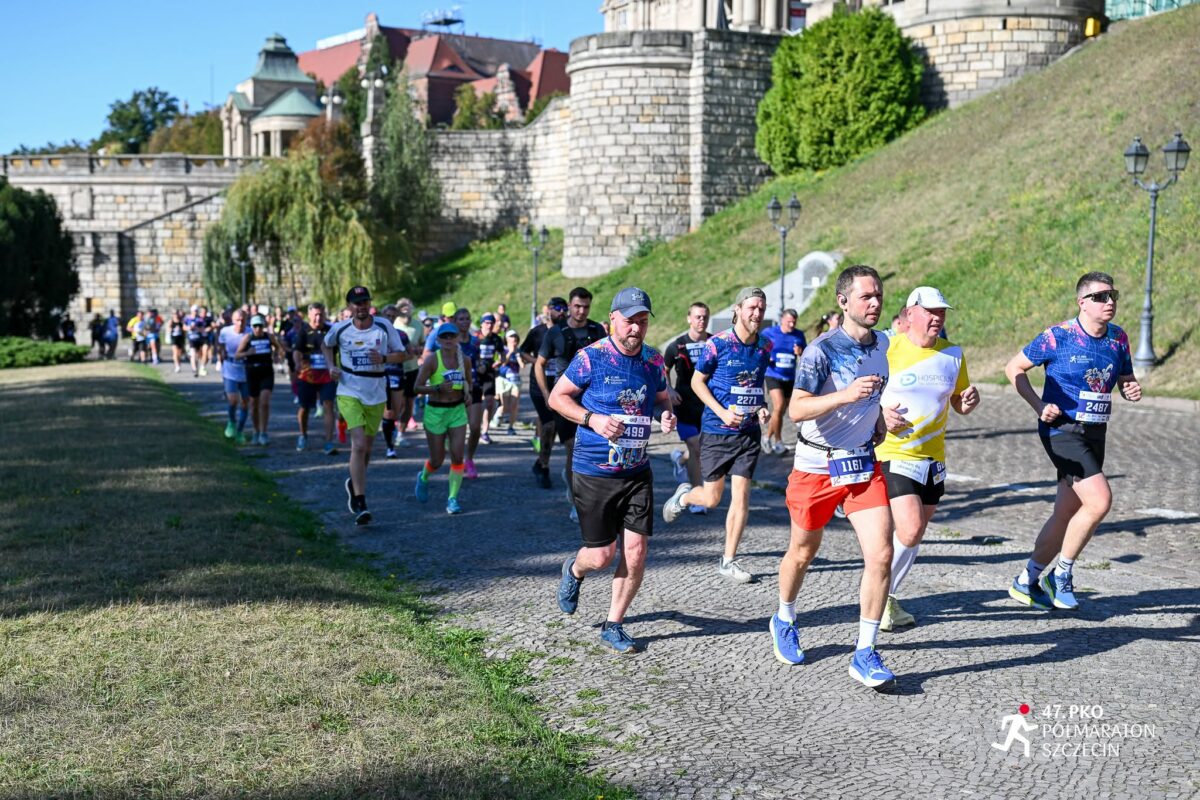 Grupa biegaczy uczestniczących w ulicznym półmaratonie. Na pierwszym planie kilkunastu dorosłych mężczyzn i kobiet biegnie po szerokiej, brukowanej alejce w słoneczny dzień. Większość ma na sobie sportowe koszulki techniczne w kolorach niebieskim, białym lub żółtym, spodenki oraz buty do biegania. Na koszulkach i biodrach widać przypięte numery startowe. Po prawej stronie u dołu widać biały logotyp z sylwetką biegacza oraz napis „47. PKO Półmaraton Szczecin”. W tle za biegaczami biegnie większa grupa uczestników. Po lewej i z tyłu znajdują się kamienne mury, schody i zabudowania o historycznym charakterze, na wzniesieniu porośniętym trawą i krzewami. Pogoda jest bezchmurna, trawa częściowo wyschnięta, a oświetlenie wskazuje na jasny, letni dzień.