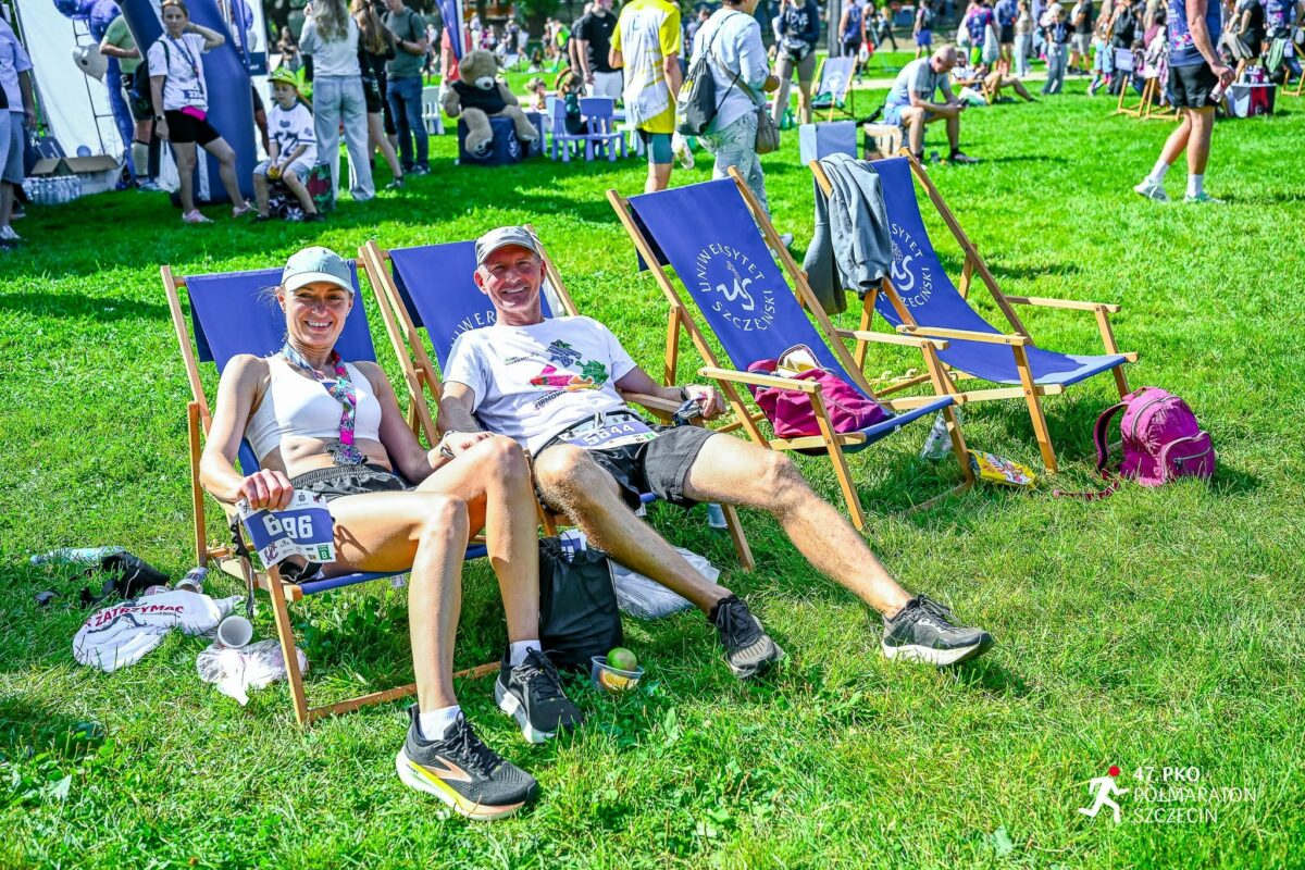 Dwoje dorosłych ludzi na leżakach na trawie podczas imprezy sportowej. Z przodu siedzi kobieta o jasnej karnacji w białym topie sportowym, krótkich jasnych spodenkach, jasnej czapce z daszkiem i czarno-białych butach do biegania; ma na szyi kolorowy medal i przypięty numer startowy 696 na pasku w pasie. Obok niej siedzi mężczyzna o jasnej karnacji w białej koszulce z kolorowym nadrukiem, czarnych krótkich spodenkach i czarnych butach do biegania; na biodrach ma przypięty numer startowy, ale jest częściowo przysłonięty i nieczytelny. Oboje uśmiechają się i odpoczywają na niebieskich materiałowych leżakach z drewnianą ramą; na oparciach leżaków widoczny jest biały napis „Uniwersytet Szczeciński” oraz okrągłe logo z literami „US”. Na trawie obok nich leżą torby, plastikowe butelki, jabłko i kilka drobnych przedmiotów; z lewej strony na ziemi znajdują się zwinięte koszulki i kubek. W tle, na dużej zielonej polanie, widać wiele innych osób – część stoi, część siedzi na leżakach lub ławkach – oraz kilka namiotów i dmuchanych konstrukcji, typowych dla biegowych stref finiszera. Po prawej stronie widać jeszcze kilka pustych leżaków z tym samym logo oraz różowy plecak na trawie. W prawym dolnym rogu zdjęcia znajduje się białe logo biegu z sylwetką biegacza i napisem „47. PKO PÓŁMARATON SZCZECIN”.