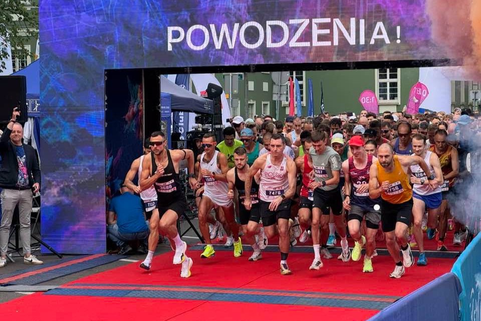 Start biegu ulicznego z dużą grupą zawodników ruszających z czerwonego dywanu spod bramy startowej. Na górze bramy widoczny jest duży napis „POWODZENIA!” na tle kolorowej, niebiesko‑fioletowej grafiki. Na pierwszym planie kilkunastu biegaczy jest w pierwszych krokach po sygnale startu, mają pochylone sylwetki i energicznie ruszają do przodu. Większość to dorośli mężczyźni w sportowych koszulkach bez rękawów lub lekkich T‑shirtach, w spodenkach do połowy uda i butach do biegania w jaskrawych kolorach, takich jak biel, żółć, limonka, czerwień czy błękit. Środkowy zawodnik ma białą koszulkę z napisem „POLSKA” i numer startowy przypięty do pasa, obok niego biegną inni z numerami na pasach lub koszulkach. Po lewej stronie, tuż przy bramie, stoi mężczyzna w jasnych spodniach i kurtce, z ręką uniesioną w górę, jakby dawał sygnał startu. Za pierwszą linią widać gęsty tłum kolejnych biegaczy, ustawionych ciasno jeden za drugim, również w kolorowych strojach sportowych. W tle po bokach pojawiają się namioty organizatorów, maszty z flagami, różowe i inne kolorowe banery oraz fragment zielonego budynku z oknami. Po prawej i lewej stronie przy starcie obecne są elementy infrastruktury biegu, takie jak barierki i maty pomiarowe na podłożu, a przy prawej krawędzi widoczny jest dym lub kolorowa mgła dodająca efektu startowi.