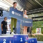 Stojące na scenie na podium dwie kobiety – uczestniczki 46. PKO Półmaratonu Szczecin, trzymające granatowe plansze z logiem Klubu Absolwentów Uniwersytetu Szczecińskiego i Centrum Badań Strukturalno-Funkcjonalnych Człowieka Uniwersytetu Szczecińskiego