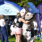 Stojąca tyłem kobieta tuli dużą uniwersytecką maskotkę – Uszatkę – sowę w granatowym birecie z białym okrągłym logiem Uniwersytetu Szczecińskiego. W tle ludzie i granatowo-biały, półokrągły, dmuchany namiot z logami uczelni.