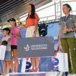Stojące na scenie na podium trzy kobiety – uczestniczki 46. PKO Półmaratonu Szczecin, trzymające puchary i papierowe torby z upominkami.