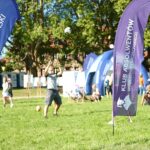 Stojące na trawie flagi (windery) – po lewej granatowy z białym logiem Uniwersytetu Szczecińskiego, po prawej fioletowy z białym logiem Klubu Absolwentów Uniwersytetu Szczecińskiego. W tle mężczyzna odbijający piłkę i ludzie siedzący na leżakach.