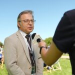 Stojący przodem Prorektor ds. Finansów – dr hab. Jacek Batóg, prof. US (ujęcie do pasa) udzielający wywiadu stojącemu tyłem dziennikarzowi, który trzyma mikrofon. W tle stojące na trawie leżaki z logiem Uniwersytetu Szczecińskiego.