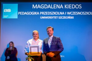 Kobieta i mężczyzna stojący na tle niebieskiego ekranu podczas uroczystości wręczenia nagrody. Kobieta po lewej stronie trzyma w rękach prostokątne wyróżnienie w ramce oraz pojedynczą różę z liśćmi. Ma jasną skórę, blond włosy związane w kucyk, ubrana jest w białą koszulę. Obok niej stoi mężczyzna o jasnej skórze, w granatowym garniturze, białej koszuli i jasnym krawacie we wzór, trzyma dłonie splecione przed sobą. W tle, po lewej, siedzi kolejna osoba – kobieta o jasnej skórze i długich blond włosach, ubrana w ciemny żakiet. Na dużym ekranie za nimi widoczny jest biały napis na niebieskim tle