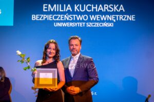 Dwoje ludzi stojących na scenie na tle niebieskiego ekranu z napisem. Po lewej stronie stoi młoda kobieta o jasnej cerze i falowanych, brązowych włosach do ramion, ubrana w elegancką, czarną, bezramiączkową suknię. W jednej ręce trzyma pojedynczą białą różę z zielonymi liśćmi, a w drugiej prostokątne, oprawione w ramę wyróżnienie lub dyplom. Uśmiecha się lekko w stronę aparatu. Obok niej, po prawej, stoi mężczyzna o jasnej cerze i krótkich, zaczesanych do tyłu włosach z lekkim siwieniem. Ma krótki zarost. Ubrany jest w ciemny garnitur, białą koszulę i jasny krawat we wzór. Trzyma złożone dłonie przed sobą i patrzy w stronę aparatu. Tło stanowi duży, podświetlony na niebiesko ekran. Na górze ekranu widoczny jest biały napis