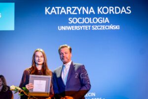 Młoda kobieta i mężczyzna stoją razem na tle niebieskiego ekranu z napisem. Kobieta ma jasną karnację, długie proste włosy w odcieniu ciemnego blondu lub jasnego brązu, okulary w cienkich oprawkach i ciemne ubranie; w dłoniach trzyma prostokątną, oprawioną w ramkę kartę lub dyplom oraz pojedynczą białą różę z zielonymi liśćmi. Obok niej stoi mężczyzna w średnim wieku o jasnej karnacji, krótkich zaczesanych do tyłu włosach w kolorze ciemny blond z siwizną i krótkim zarostem; ma na sobie ciemnogranatowy garnitur, białą koszulę i jasny krawat z drobnym wzorem. Tło stanowi duży, jednolicie niebieski ekran z białym tekstem, na którym u góry widnieje napis