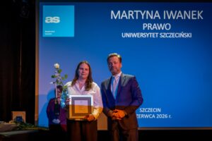 Młoda kobieta i mężczyzna stoją na tle dużego niebieskiego ekranu podczas uroczystości. Kobieta o jasnej karnacji i długich, brązowych włosach ma na sobie białą, zapinaną koszulę i ciemne spodnie. Trzyma w rękach prostokątną, oprawioną w ramę dyplomową tablicę oraz pojedynczą białą różę z długą łodygą i liśćmi. Obok niej stoi mężczyzna o jasnej karnacji, krótkich zaczesanych do tyłu włosach i brodzie; ubrany jest w ciemny garnitur, jasną koszulę i jasny krawat we wzór. Ma splecione dłonie przed sobą i patrzy w stronę aparatu. W tle, po lewej stronie, widoczna jest kobieta w bordowej marynarce i czarnej bluzce, stoi nieco w cieniu. Na stole obok leżą rozłożone dokumenty lub książki oraz kilka długich łodyg z kwiatami. Tło z prawej strony wypełnia ekran z wyświetlonym niebieskim slajdem.