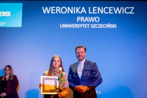 Młoda kobieta i mężczyzna stoją na scenie na tle niebieskiego ekranu z napisem. Kobieta po lewej stronie ma jasną karnację, proste, długie, jasnobrązowe włosy i delikatny makijaż. Ubrana jest w jasny golf i trzyma w rękach prostokątną, oprawioną w ramkę dyplomową tablicę oraz pojedynczą białą różę z zielonymi liśćmi. Mężczyzna po prawej stronie ma średnią karnację, krótkie ciemne włosy z siwizną i krótki zarost. Ubrany jest w ciemny garnitur, białą koszulę i jasny krawat ze wzorem. Stoi z rękami złożonymi z przodu.
