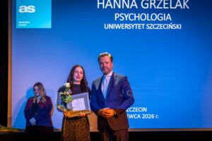 Scena wręczenia nagrody na tle dużego niebieskiego ekranu. Na środku stoją obok siebie dwie osoby: po lewej młoda kobieta o jasnej skórze i długich, ciemnych włosach, ubrana w ciemną bluzkę z krótkim rękawem i ciemną spódnicę w drobne jasne kropki, trzymająca w jednej dłoni białą różę z zielonymi liśćmi, a w drugiej prostokątne ujęte w ramkę dyplomowe wyróżnienie. Po prawej stoi mężczyzna o jasnej skórze, krótko zaczesanych do tyłu włosach z siwizną, w ciemnym garniturze, jasnej koszuli i krawacie w jasny wzór; obie dłonie ma złożone przed sobą. W głębi po lewej stronie widoczna jest jeszcze jedna kobieta o jasnej skórze i długich, jasnych włosach, w ciemnej marynarce, stojąca z rękami skrzyżowanymi przed sobą. Na górze niebieskiego ekranu wyświetlony jest biały napis