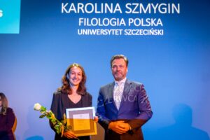 Młoda kobieta o jasnej karnacji z długimi brązowymi włosami trzymająca dyplom w ramce i pojedynczą białą różę. Stoi na scenie obok mężczyzny o jasnej karnacji w eleganckim garniturze w kolorze granatowym, z białą koszulą i jasnym krawatem we wzór. Kobieta ma na sobie ciemny żakiet i bluzkę z dekoltem w serek, uśmiecha się lekko w stronę aparatu. Dyplom ma jasną ramę i beżowy środek z nadrukowanym tekstem i herbem u góry, treść nie jest czytelna. Biała róża ma długą zieloną łodygę z liśćmi i jest owinięta u dołu wstążką. Tło jest gładkie, w intensywnym niebieskim kolorze, oświetlone światłem scenicznym. Nad nimi widoczny jest jasny napis: „KAROLINA SZMYGIN” w największym rozmiarze, pod spodem mniejszymi literami „FILOLOGIA POLSKA” oraz „UNIWERSYTET SZCZECIŃSKI”. W lewym górnym rogu znajduje się jasny prostokąt z fragmentem białego znaku graficznego. Po lewej stronie w tle siedzi jeszcze jedna osoba w ciemnym stroju, częściowo widoczna, mniej oświetlona. Cała scena ma charakter uroczystości wręczenia wyróżnienia lub nagrody.