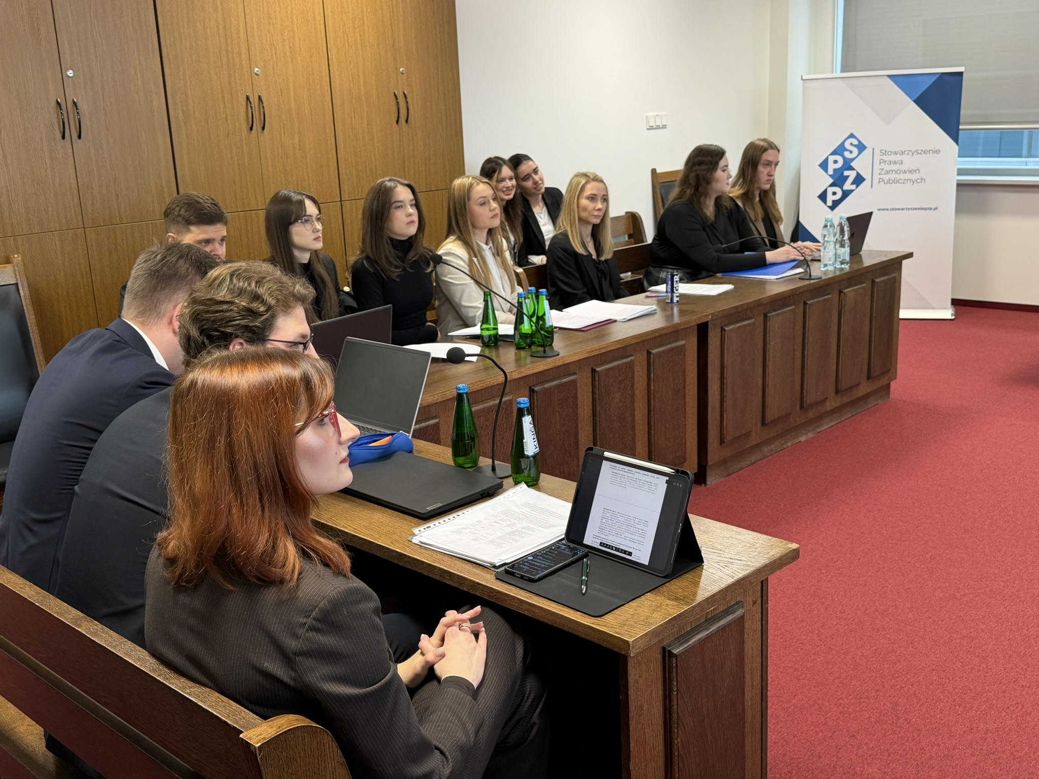 Pomieszczenie do symulacji rozprawy sądowej. Za drewnianymi zabudowanymi stołami siedzi młodzież – przed niektórymi stoją laptopy lub tablety.