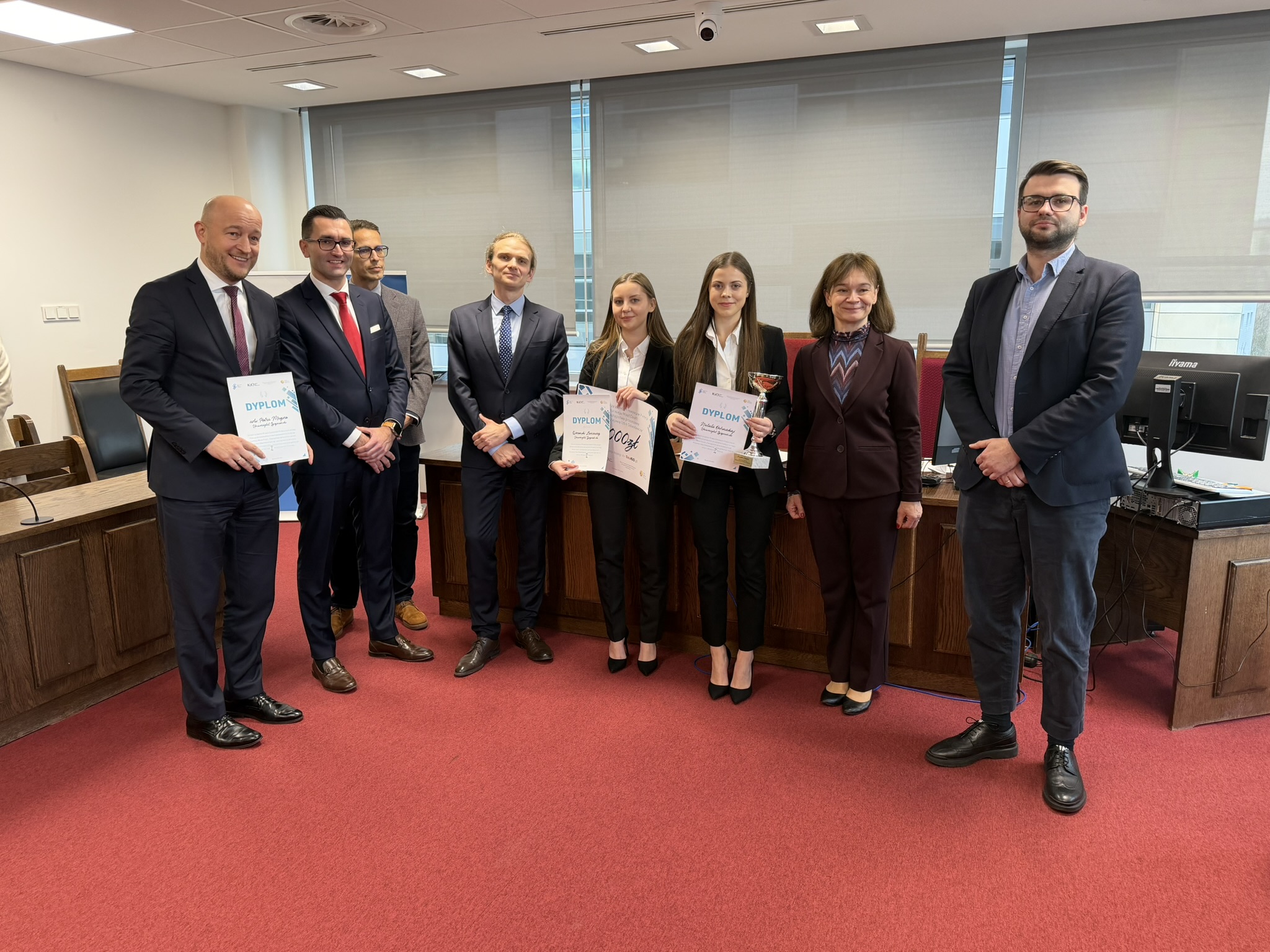 Pomieszczenie do symulacji rozprawy sądowej. Na bordowej wykładzinie stoją jurorzy z dwiema studentkami – laureatkami konkursu, które trzymają puchar, dypolomy i symboliczny czeka na 8 tysięcy złotych.