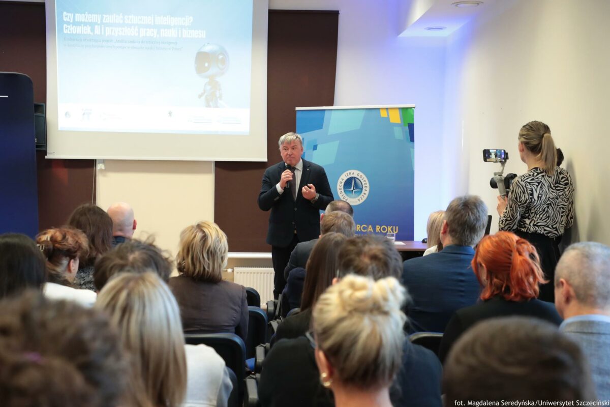 Prorektor ds. nauki Uniwersytetu Szczecińskiego otwierający konferencję o AI. Za nim rollup Północnej Izby Gospodarczej i ekran z prezentacją. Na pierwszym planie słuchacze tyłem do kadru.