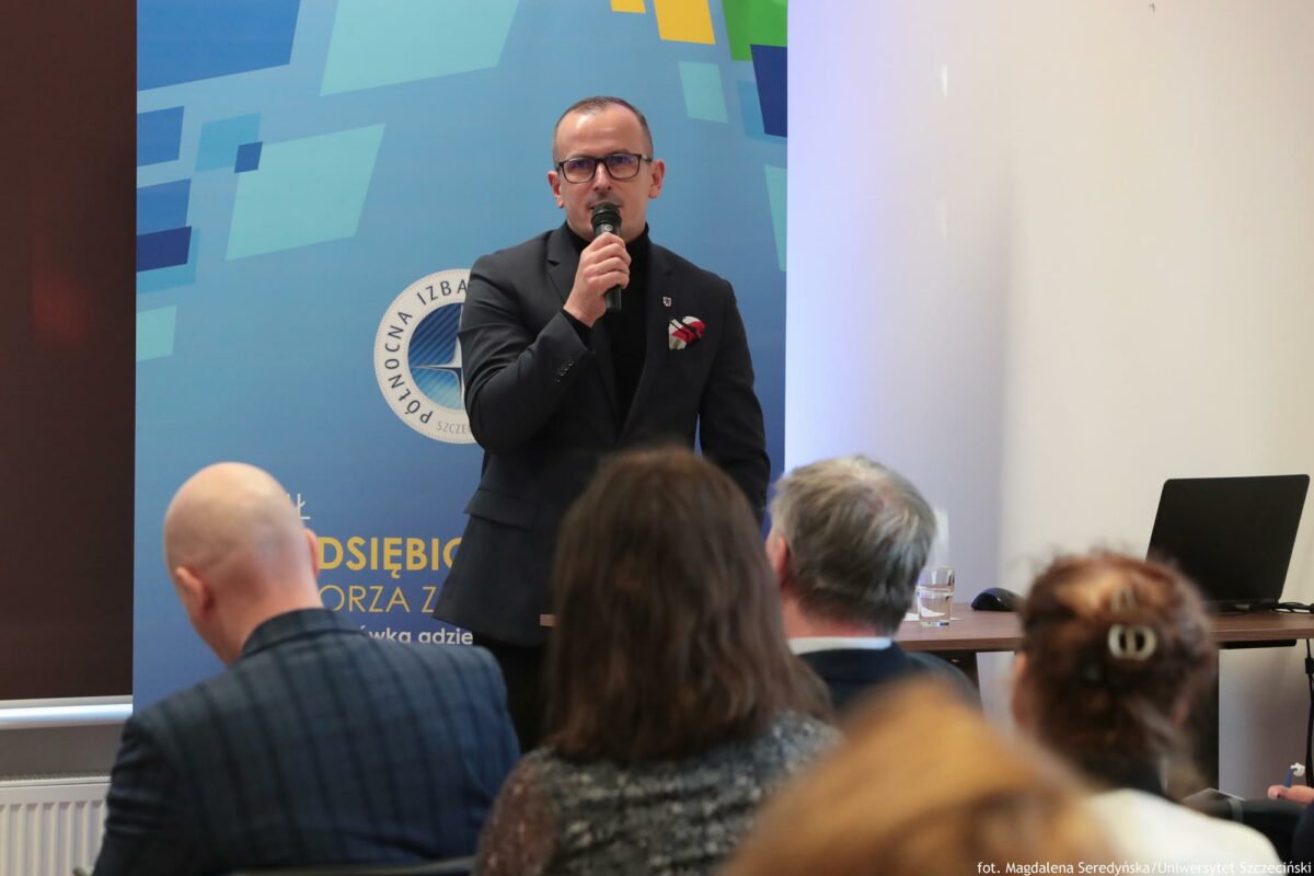 Prelegent. Przed nimi biurko z laptopem. Za nim rollup Północnej Izby Gospodarczej. Na pierwszym planie słuchacze tyłem do kadru.