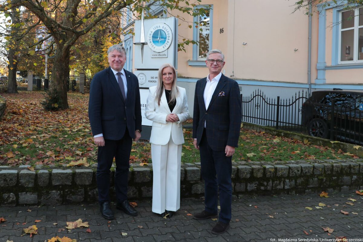 Troje uczestników konferencji o AI (kobieta w środku) przed murkiem. W tle po prawej zabytkowa willa i logo Północnej Izby Gospodarczej. Po lewej drzewo, na trawniku żółte liście.