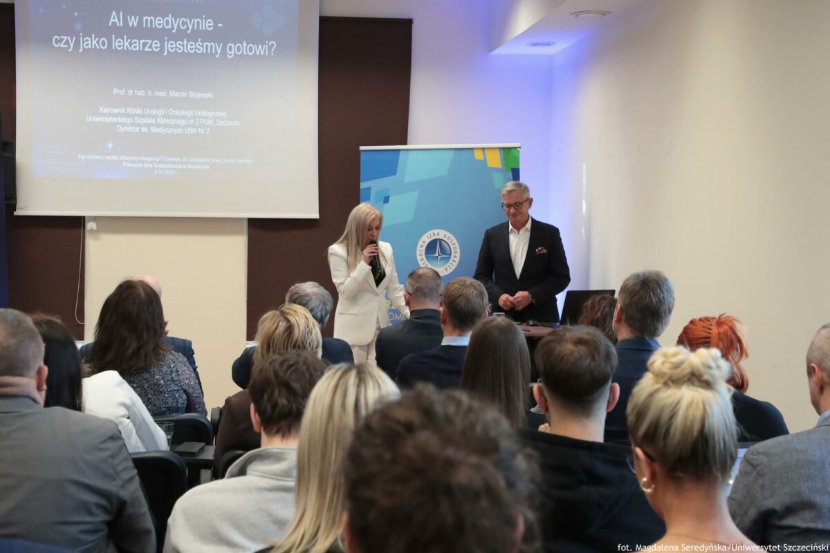 Po prawej prelegent i prowadząca. Przed nimi biurko z laptopem. Po lewej ekran z prezentacją i tytułem: „AI w medycynie”. Na pierwszym planie słuchacze tyłem do kadru.
