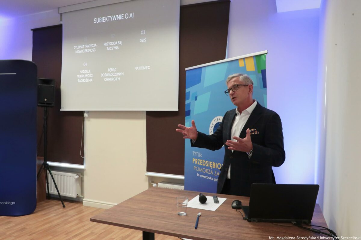 Po prawej tłumaczący coś mężczyzna. Przed nim biurko z laptopem. Po lewej ekran z prezentacją i tytułem: „Subiektywnie o AI”.