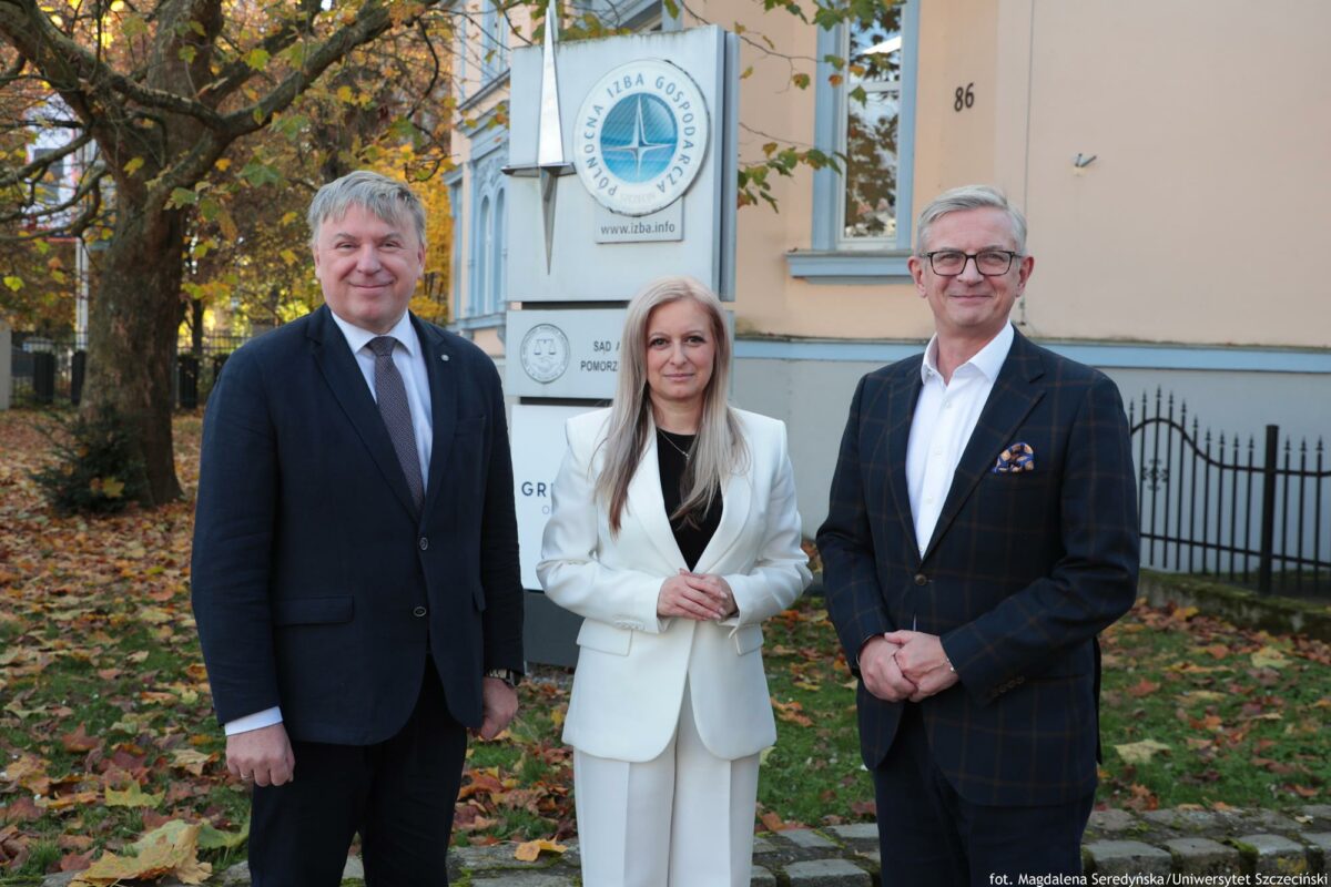 Troje uczestników konferencji o AI (kobieta w środku) przed murkiem. W tle po prawej zabytkowa willa i logo Północnej Izby Gospodarczej. Po lewej drzewo, na trawniku żółte liście.