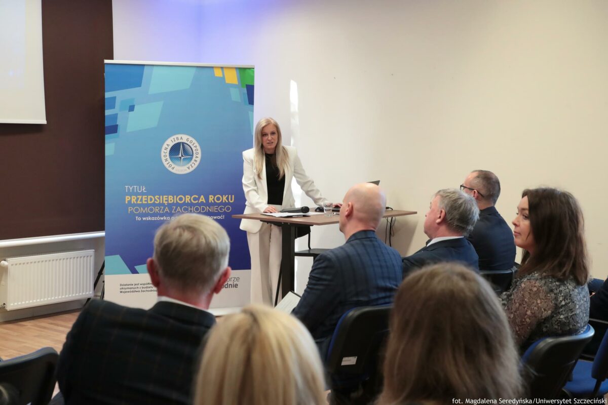 Prelegentka. Przed nią biurko z laptopem. Za nią rollup Północnej Izby Gospodarczej. Na pierwszym planie słuchacze bokiem lub tyłem do kadru.