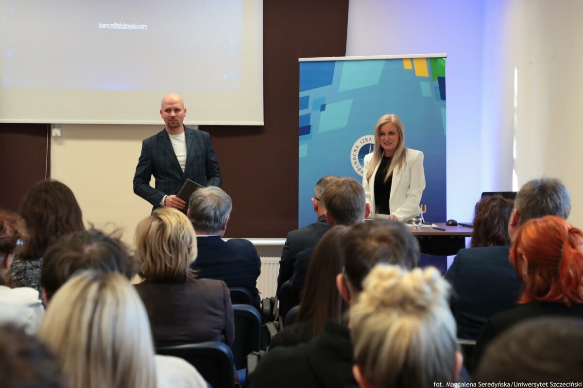 Prelegent i prowadząca. Przed nią biurko z laptopem. Za nią rollup Północnej Izby Gospodarczej. Za mężczyzną ekran z prezentacją. Na pierwszym planie słuchacze tyłem do kadru.