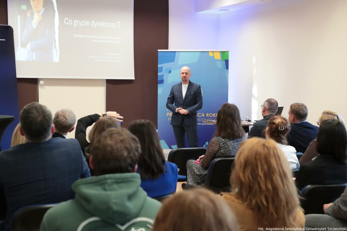 Prelegent. Obok niego biurko z laptopem. Za nim rollup Północnej Izby Gospodarczej i ekran z prezentacją i tytułem: „Co gryzie dyrektora IT?”. Na pierwszym planie słuchacze tyłem do kadru.