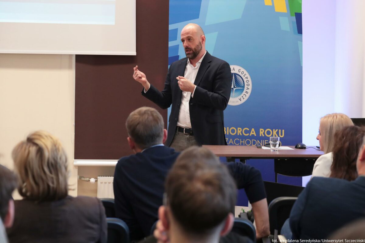 Tłumaczący coś prelegent. Przed nim biurko z laptopem. Za nim rollup Północnej Izby Gospodarczej. Na pierwszym planie słuchacze tyłem do kadru.